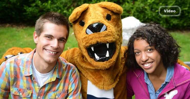 The lion sits with two smiling students on a campus bench.